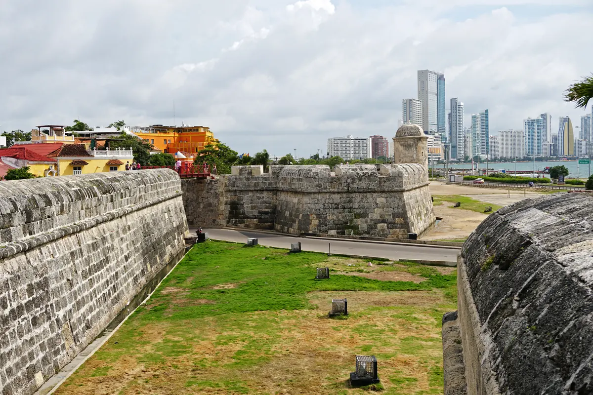 A stone bastion and long city wall with the Bocagrande skyline in the distance under a cloudy sky