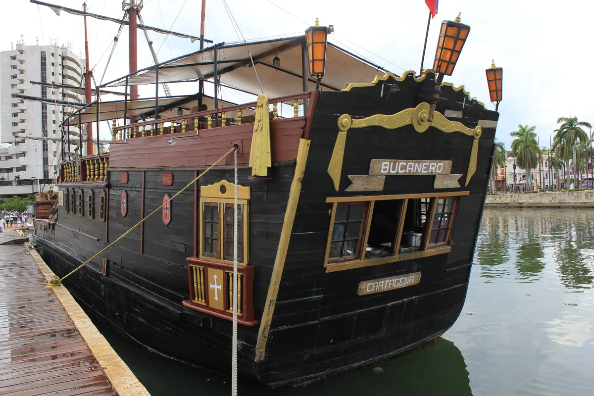 The stern of a black pirate-style tour ship with gold carving, lanterns and a name board, moored at a wooden pier.