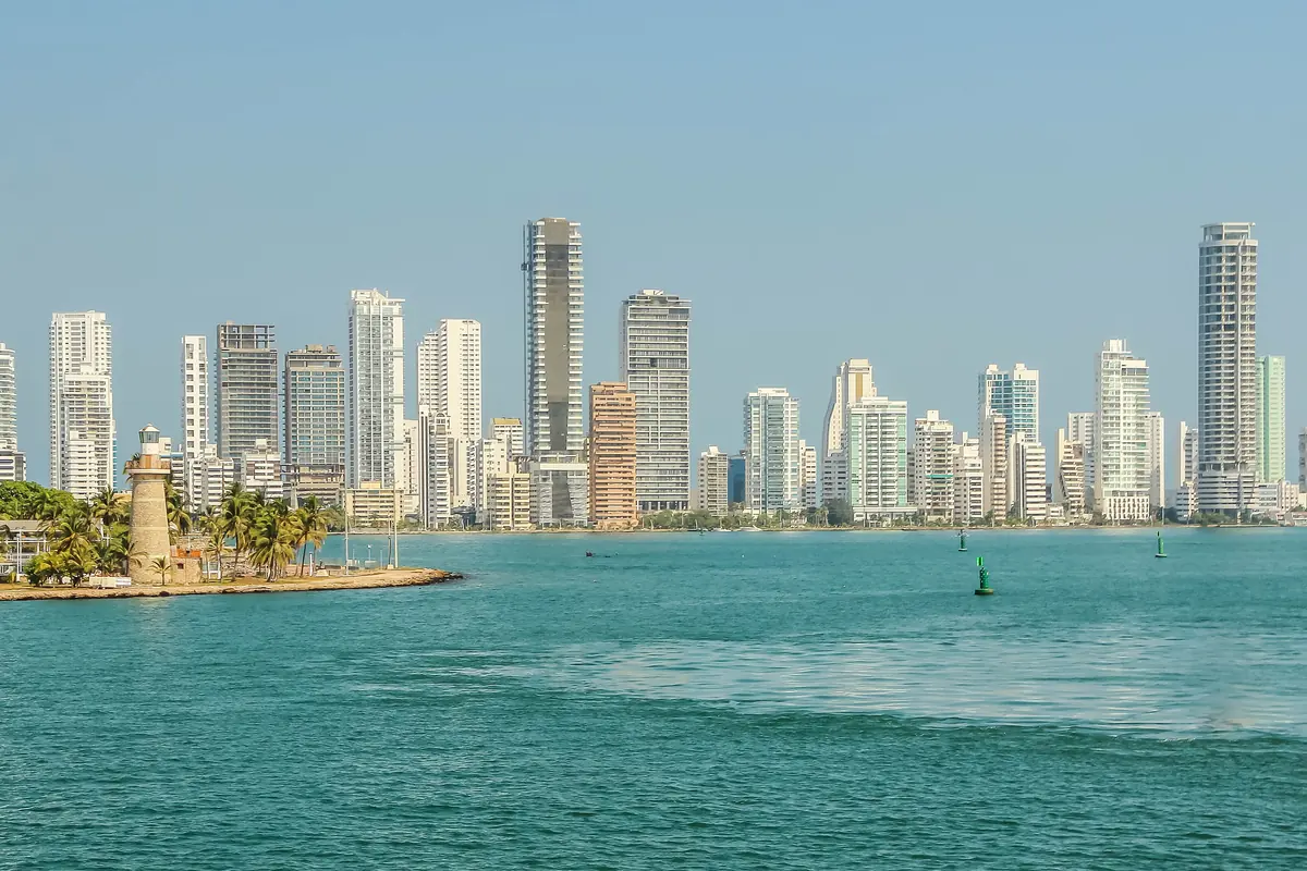 A white lighthouse on a green point with the tall apartment towers of Bocagrande rising behind it across turquoise water.