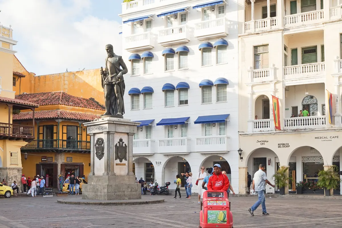 A bronze statue on a stone plinth in a plaza lined with white colonial buildings with balconies and arches