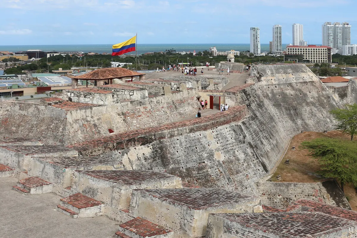 The grey stone ramparts of the Castillo San Felipe de Barajas, with a Colombian flag and the Cartagena skyline beyond.