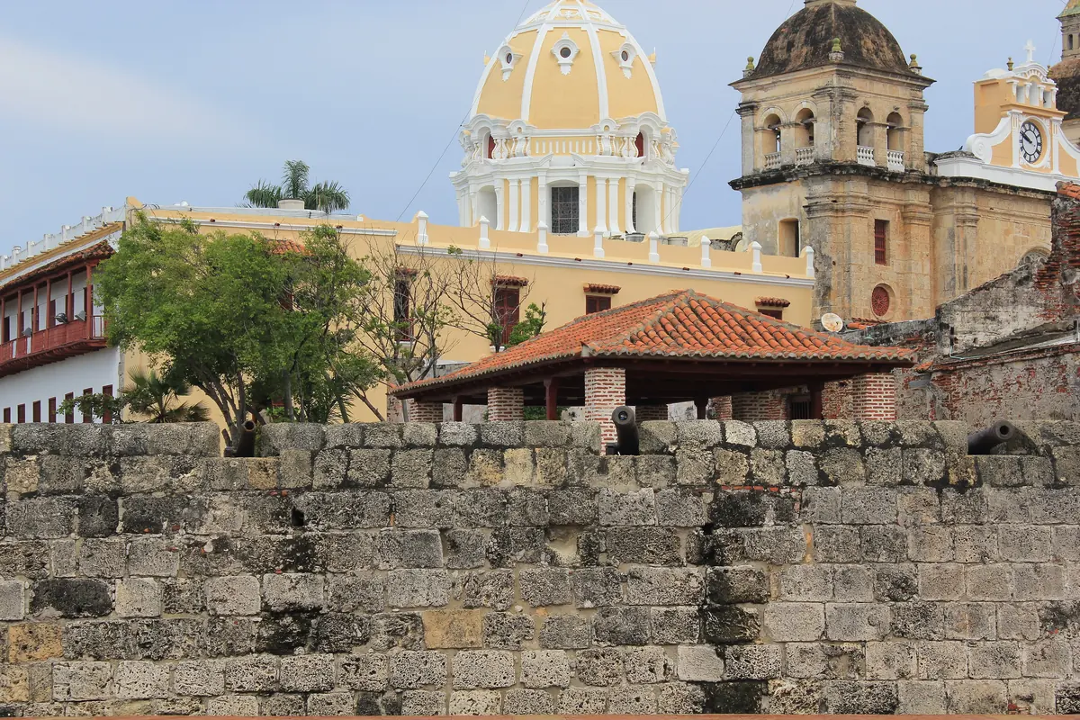A weathered stone city wall with an old cannon, and a yellow church dome and tower behind it.